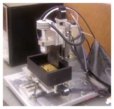 Compact milling machine with black body and silver components on table, featuring square base plate and vertical column with cutting tool spindle head.