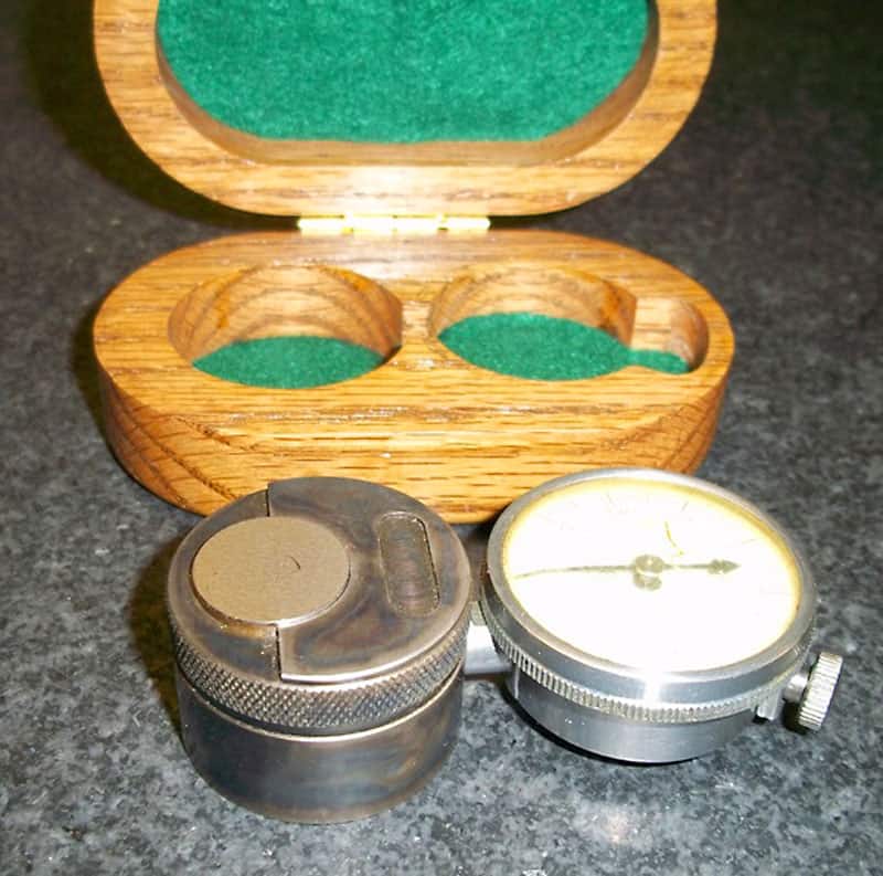 Wooden box with green felt lining containing two small metal objects on a gray surface.