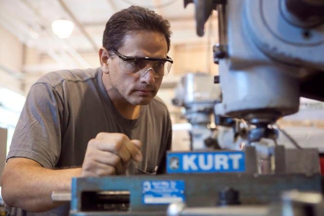 A man wearing safety glasses and a gray t-shirt stands behind a blue KURT machine.