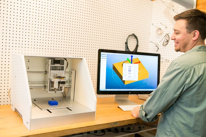 A man stands in front of a computer monitor displaying a 3D model, with an open white printer behind him against a pegboard wall background.