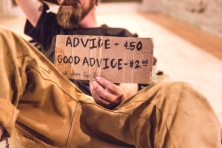 A man sitting on the ground with a cardboard sign reading "ADVICE - $.50 GOOD ADVICE - $2.00" and "BAD JOKES for free".