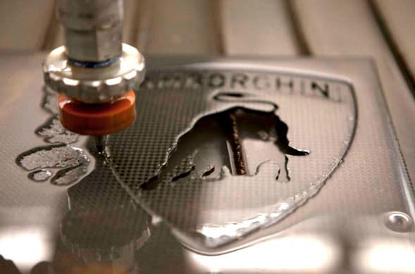 Water jet cutting tool in action, pointed at camera on top of stainless steel or aluminum metal sheet with Norghin logo featuring a bull's head and cross.
