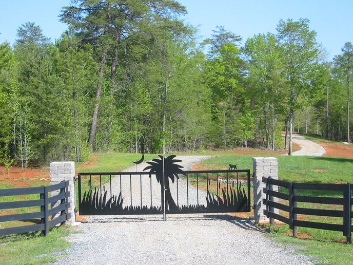 Rural entrance with black gate, palm tree cutout, and stone pillars in a serene landscape setting.