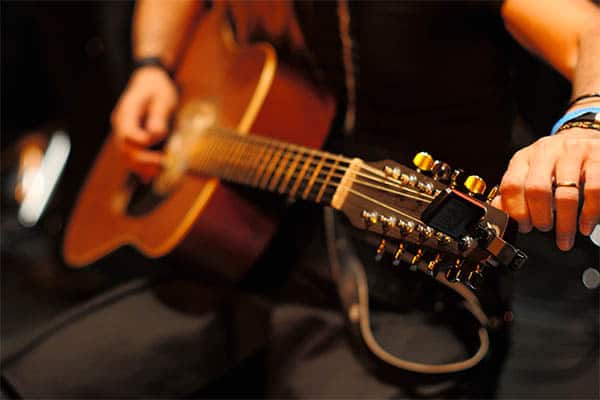 A person's hands in the background, with a guitar positioned in front, being played by someone not shown.