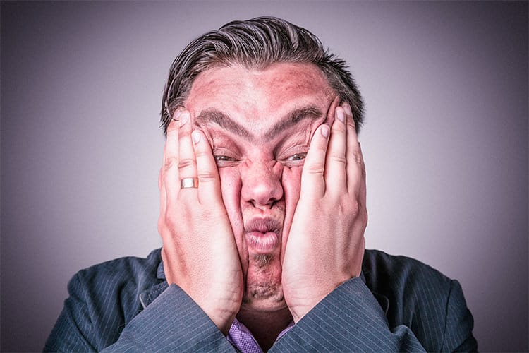 A man with his hands on his face, wearing a suit jacket and pink shirt, experiencing frustration.