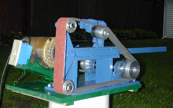 Red sanding belt grinder with metal components on a green table in a dark environment with green grass in the background.