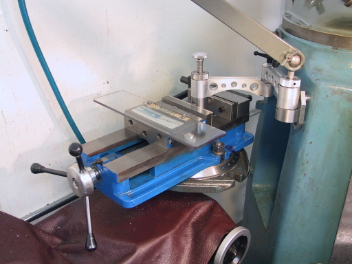 A blue benchtop vise with swing arm against a white wall, featuring a black handle and adjustable metal jaw.
