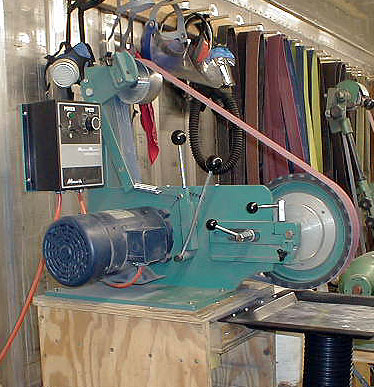 A green belt sander with black motor and red cord on a wooden table in a workshop setting.