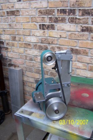 A metal bench grinder sits atop a painted metal table with a weathered brick wall backdrop.