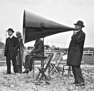 Three men in old-fashioned suits standing on a gravel surface, one holding a megaphone.