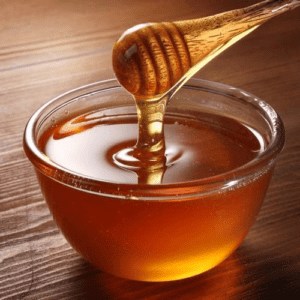 A small glass bowl containing golden honey and a wooden honey dipper with honey on the end, set against a dark wood surface.