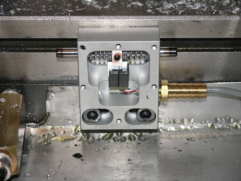 A metal component with square shape and holes, attached to smaller parts in a manufacturing context.