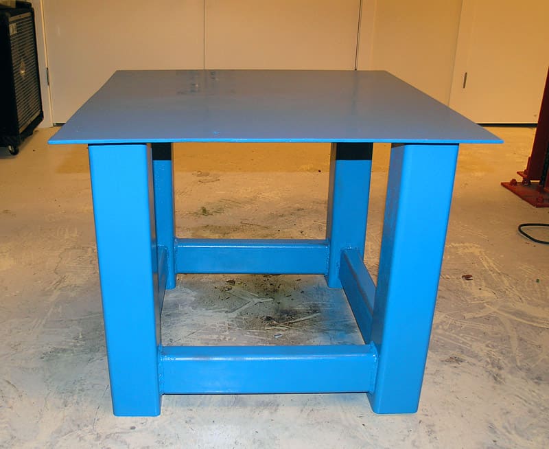 A sturdy blue metal or wood table with four square legs on a steel or aluminum surface.