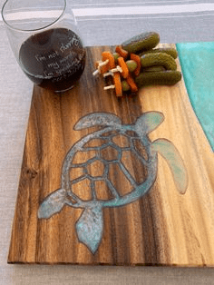 Wooden cutting board with light-colored wood grain and blue-green resin turtle design, accompanied by a glass of red wine and pickled vegetables.