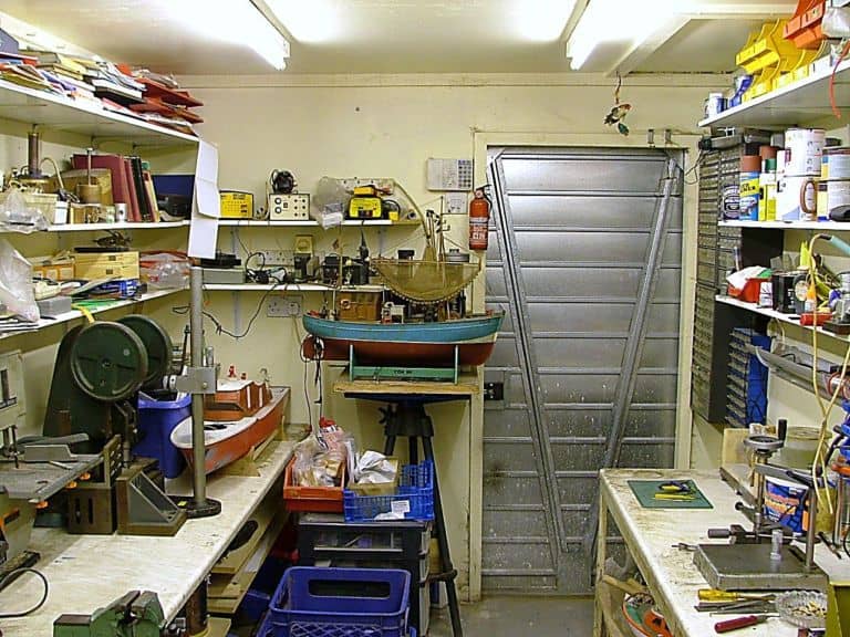 Workshop interior with shelves, workbenches, fluorescent lights, metal door, tables, power tools, hand tools, and machine parts.