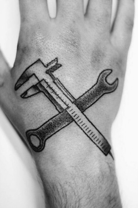 A black and white image of a hand with a tattoo of crossed wrenches and calipers on the wrist.